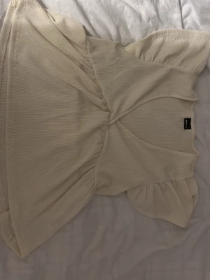Beige ribbad omlottopp från H&M - Supersöt beige omlottopp från gina med ribbad struktur och korta puffärmar. Toppen har en v-ringad urringning och är gjord i ett mjukt, stretchigt material som sitter bekvämt. Perfekt att styla med jeans eller kjol.