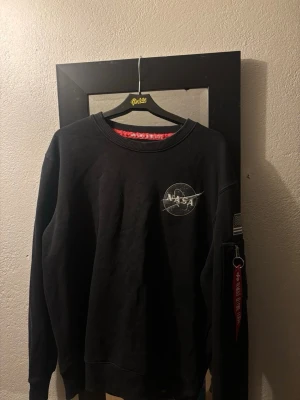 Svart NASA sweatshirt med patch - Cool svart sweatshirt med NASA-logga på bröstet och amerikansk flaggpatch på ärmen. Tröjan har rund hals, långa ärmar och en ficka med dragkedja och röd tag på ena ärmen. Perfekt för dig som gillar rymdtema och streetwear.