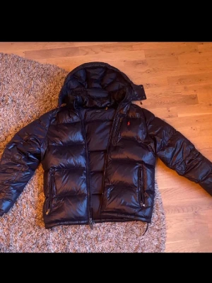 Svart dunjacka från Polo Ralph Lauren - Snygg svart dunjacka från Polo Ralph Lauren med glansig finish och stor huva. Jackan har två stora framfickor med dragkedja och diskret röd logga på bröstet. Perfekt för kalla dagar och riktigt skön passform.