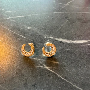 Guldtonade örhängen med stenar - Säljer ett par små, halvcirkelformade örhängen i guldton med gnistrande vita stenar. Örhängena har en modern och stilren design som ger en lyxig känsla. Perfekta för dig som gillar detaljer som sticker ut.