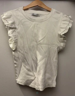 Vit ribbad topp med volangärmar Zara - Supersöt vit ribbad topp från Zara med volangdetaljer på ärmarna. sopassad rund halsringning och croppad passform. Perfekt att styla med jeans eller kjol för en fräsch look. Denna topp är använd en till två gånger och är därför i ett mycket bra skick