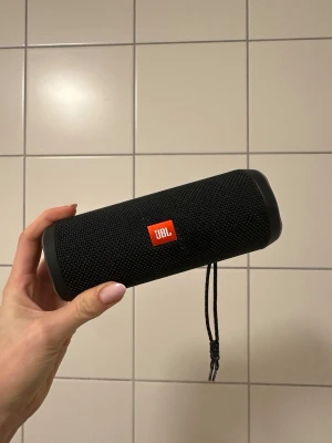 JBL svart bärbar Bluetooth-högtalare - Snygg och kompakt bärbar högtalare från JBL i svart. Cylindrisk form med tåligt tygmaterial och gummidetaljer. Utrustad med praktisk handledsrem och tydliga knappar för volym och uppspelning. Perfekt för att ta med musiken överallt.