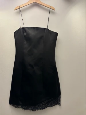 Svart satin klänning med spetskant - Stilren svart kort klänning i satin med tunna axelband och rak urringning. Klänningen har en asymmetrisk nederkant dekorerad med svart spets som ger en elegant touch. Perfekt om du ska på en finare tillställning! Den är ny med prislapp och har alltså inte använts på. Nytt skick!