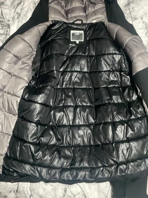 Svart dunjacka med huva från Jack and Jones - Snygg svart dunjacka från Jack and jones med quiltad insida och grå detaljer. Jackan har huva, dragkedja framtill och två sidofickor med dragkedja. Justerbara muddar med kardborreband för extra komfort. Perfekt för kalla dagar.