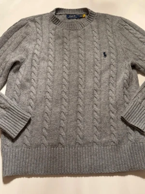 Grå kabelstickad tröja Polo Ralph Lauren - Säljer en grå kabelstickad tröja från Polo Ralph Lauren. Tröjan har rund halsringning, ribbade muddar och nederkant samt den klassiska broderade loggan i marinblått på bröstet. Perfekt för dig som gillar stilrena och tidlösa plagg.