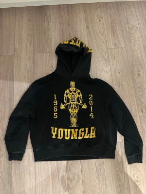 Svart YoungLA hoodie med tryck - Säljer en svart hoodie från YoungLA med stort gult tryck av en bodybuilder goldsgym och texten '1965-2014' samt 'YOUNGLA' på framsidan. Huvan har också gult tryck och på ryggen finns en liten gul logga. Hoodien har en klassisk passform och känguruficka.