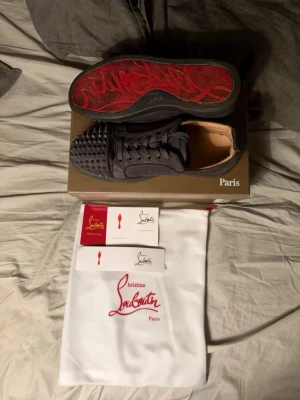 Christian Louboutin svarta sneakers med nitar - Grymma svarta sneakers från Christian Louboutin med ikonisk röd sula och coola nitar framtill. Skorna har snörning och är tillverkade i skinn med detaljer i mocka. Kommer med originalkartong, dustbag och äkthetskort. Perfekta för dig som vill sticka ut.