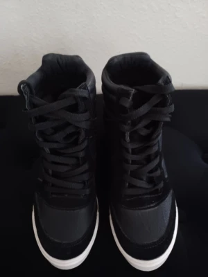 Svarta high top sneakers med vit sula - Säljer ett par svarta high top sneakers med vit sula och snörning. Skorna har en cool, sportig look med vadderad kant och detaljer i syntetmaterial och mockaimitation. Perfekta för dig som gillar streetstyle och vill ha något bekvämt och snyggt. Klackhöjd: 9 cm. Använda endast 1 gång.