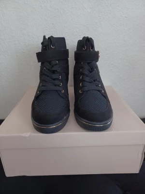 Svarta höga sneakers med snörning - Snygga svarta sneakers med högt skaft, snörning och kardborreband. Skorna har detaljer i guld och är tillverkade i en mix av mesh och mockaimitation. Perfekta för dig som gillar streetstyle och vill ha något som sticker ut. Klackhöjd: 7 cm. Orginalkartong: 29×26×11. Endast provade.