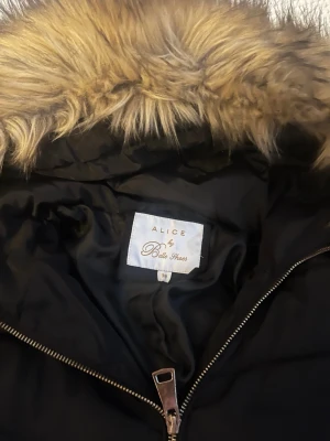 Svart dunjacka med fuskpäls från Alice by belle shoes - Svart dunjacka från Alice by Bella Shoes med stor luva kantad av beige fuskpäls. Jackan har quiltad design, dragkedja framtill och två fickor med dragkedjor. Nypris 1299kr