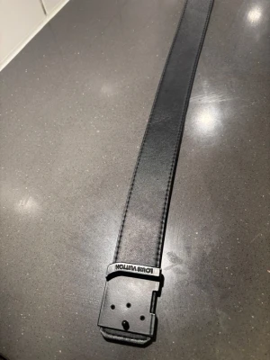 Svart läderbälte - Snyggt svart bälte i äkta läder med diskret präglad logotyp på spännet. Klassisk och stilren design som passar till jeans eller kostymbyxor. Tillverkat i Spanien och har tydliga sömmar längs kanterna.