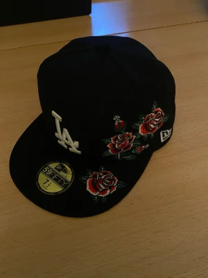 Svart Firted LA-keps med rosbrodyr från New Era 7 1/6 - ( STORLEK 7 1/6  ca 60cm ) Snygg svart keps från New Era med LA-logga framtill och broderade röda rosor på sidan. Klassisk 59FIFTY-modell med platt skärm och broderade detaljer. Perfekt för dig som vill sticka ut med en unik accessoar.