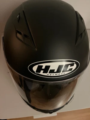 Svart MC-hjälm från HJC - Säljer en svart integralhjälm från HJC med tydlig logga framtill. Hjälmen har en slät, rundad form och är utrustad med ventilationsöppningar upptill samt ett klart visir. 