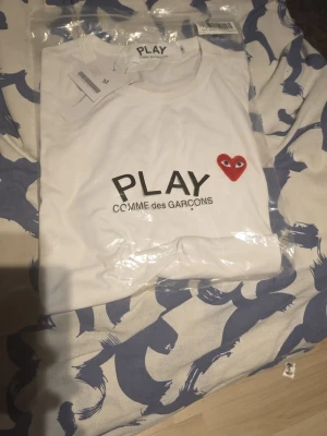 Vit Comme des Garçons PLAY t-shirt - Säljer en vit t-shirt från Comme des Garçons PLAY med det ikoniska röda hjärtat med ögon på bröstet och ett stort hjärta tryckt längst ner. Klassisk rund hals och korta ärmar. T-shirten är i mjuk bomull och har en clean, stilren look.