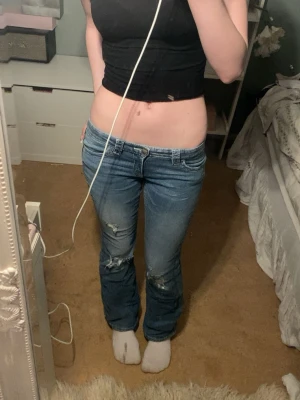 Blå lågmidjade bootcut jeans - Säljer ett par blå lågmidjade bootcut jeans med slitningar på knäna och snygga detaljerade bakfickor. Jeansen har klassisk femficksdesign och är tillverkade i jeansmaterial med en skön passform.