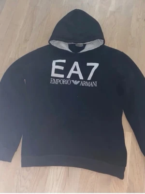 Svart Emporio Armani Hoodie - Svart hoodie från EA7 Emporio Armani med stor vit logga broderad på bröstet. Klassisk huva med ljus insida och långa ärmar. Perfekt för dig som gillar stilrena och exklusiva streetwear-plagg.