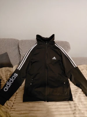 Svart Adidas hoodie med dragkedja - Säljer en svart hoodie från Adidas med klassiska vita ränder längs ärmarna och Adidas-logga på bröstet och ärmen. Tröjan har huva, hel dragkedja framtill och två fickor med dragkedja. Perfekt för dig som gillar sportig stil.