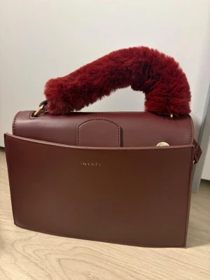 Vinröd handväska från Inyati - Säljer en stilren vinröd handväska från Inyati med ett unikt handtag i mjuk, fluffig pälsimitation. Väskan har en elegant, boxig form och guldfärgade detaljer. Perfekt för dig som vill ha något som sticker ut och känns lyxigt.