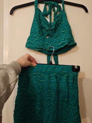 Turkost/grönt set med vågmönster - Säljer ett turkost tvådelat set i meshmaterial med vågigt mönster. Toppen har knytband i nacken och en liten cut-out framtill. Kjolen är matchande och har resår i midjan. Perfekt för sommaren och festivaler!