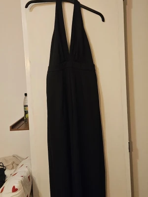 Svart halterneck jumpsuit - Snygg svart jumpsuit med djup v-ringning och halterneck. Vida ben och markerad midja ger en stilren och trendig look. Perfekt för dig som vill sticka ut med en enkel men elegant outfit.