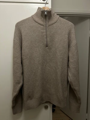 Beige stickad halvzip från Jack & Jones - Beige stickad tröja från Jack & Jones med hög krage och halvzip-dragkedja. Tröjan har ribbstickade muddar vid ärmslut och nederkant, samt en avslappnad passform. Perfekt för lager-på-lager och enkel att matcha med jeans eller chinos.