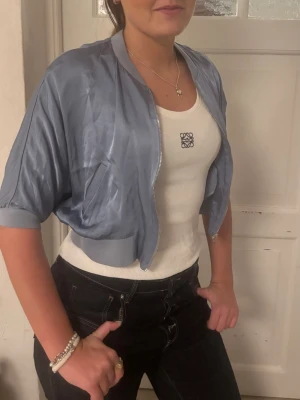 Kortämad kofta  - Snygg ljusblå bolero i satinliknande material med korta ärmar och ribbade muddar. Croppad modell som passar perfekt över linne eller klänning för en trendig lager-på-lager-look. Dragkedja framtill och avslappnad passform.