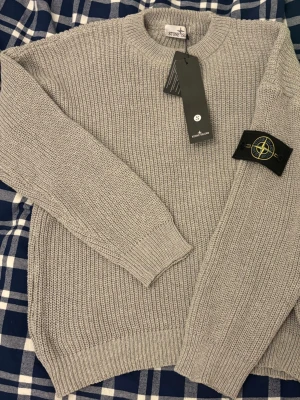 Ljusgrå stickad tröja från Stone Island - Säljer en ljusgrå stickad tröja från Stone Island med klassisk patch på ärmen. Tröjan har rund halsringning, ribbade muddar och är långärmad. Perfekt för dig som gillar stilrena och tidlösa plagg.