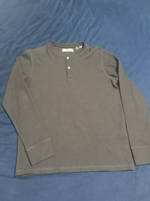 Mörkgrå långärmad henleytröja - Säljer en mörkgrå långärmad henleytröja från Polarn O. Pyret. Tröjan har rund halsringning och två knappar framtill. Tillverkad i mjukt, ribbat material som är skönt mot huden. Perfekt för lager-på-lager och enkel att matcha med olika outfits.