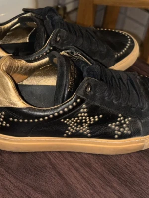 Zadig & Voltaire svarta sneakers med nitar  - Sååå snygga svarta sneakers med guldfärgade/bronsfärgade nitar i stjärnmönster på sidorna och guldfärgad insida. 100% läder/skinn. Skorna har svart snörning!! Superskick!! Passar både till fest och till vardags!! Kan skicka dem redan idag🤎🤎