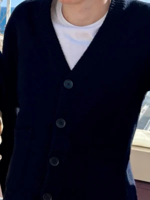 Mörkblå kofta med knappar - Snygg mörkblå kofta med stora knappar framtill och två fickor. Koftan har en djup V-ringning och ribbade muddar. Perfekt att slänga över en t-shirt för en avslappnad look.