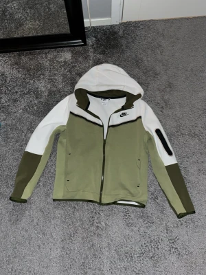 Nike hoodie i vitt och olivgrönt - Nike hoodie i vitt och olivgrönt med dragkedja framtill och huva. Snygg kontrast med mörkare gröna detaljer på ärmar och insida av huvan. Liten Nike-logga på bröstet och praktisk ficka med dragkedja på ena ärmen. Perfekt för en sportig och avslappnad stil.