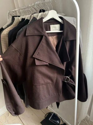 Brun ulljacka från Dagmar med bälte aviator  - Snygg mörkbrun jacka från Dagmar i ull med bred krage och bälte i midjan. Jackan har stora fickor framtill och knappar vid ärmsluten. Perfekt för dig som vill ha en stilren och trendig look. Modellen heter aviator. 