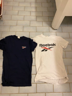 2-pack Reebok t-shirts XS - Två klassiska Reebok t-shirts i storlek XS. En marinblå med liten logga på bröstet och en vit med stor Reebok-logga framtill. Båda har rund hals och korta ärmar. Perfekta till jeans eller shorts för en sportig vibe.