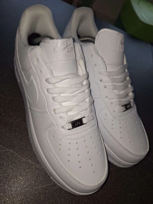 Nike Air Force 1 vita sneakers - Klassiska helvita Nike Air Force 1 sneakers med rund tå, snörning och perforerade detaljer på tån. Skorna har en platt sula och är tillverkade i syntetmaterial med Nike-logga på sidan och hälen. Perfekta för en clean och stilren look.