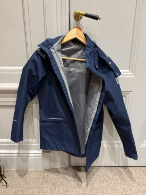 Mörkblå regnjacka med huva från Reima - Snygg mörkblå regnjacka från Name It med huva och dragkedja framtill. Jackan har två fickor med reflexdetaljer och justerbara ärmslut. Perfekt för regniga dagar och enkel att matcha med andra plagg.