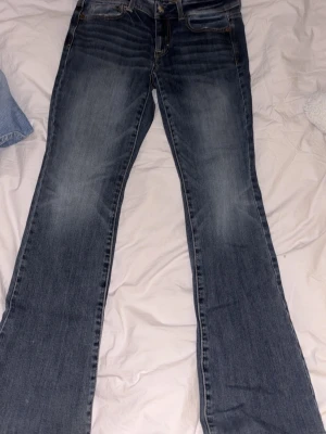 Blå bootcut jeans från American Eagle - Snygga blå bootcut jeans från American Eagle med klassisk femficksdesign och subtila slitningar framtill. Jeansen har en något utsvängd passform nedtill och är tillverkade i mjuk bomullsdenim med en skön känsla. Perfekta för en avslappnad och trendig look.