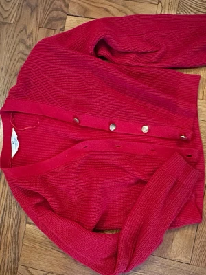 Röd ribbstickad kofta med knappar - Snygg röd ribbstickad kofta med silverfärgade knappar framtill. Koftan har en klassisk v-ringad design och långa ärmar. Perfekt att slänga över en t-shirt för en avslappnad men stilren look.