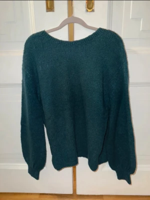 Mörkgrön stickad tröja med ballongärm - Mysig stickad tröja i mörkgrönt med rund halsringning och ballongärmar, samt öppen rygg. Tröjan har ribbade muddar vid ärmslut och nederkant, vilket ger en avslappnad men ändå snygg look. Perfekt för kyliga dagar och enkel att matcha med jeans eller kjol.