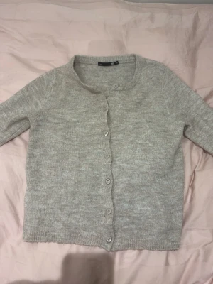 Ljusgrå kofta med rund hals från Lager 157 - Mysig ljusgrå kofta med rund halsringning och knäppning framtill. Koftan har långa ärmar och ribbade muddar vid ärmslut och nederkant. Perfekt att slänga över en topp för en enkel och stilren look. 