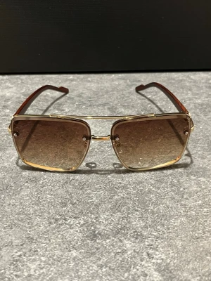 Solglasögon i hög kvalitet  - Snygga solglasögon i hög kvalitet med fyrkantiga linser och guldfärgad metallbåge. Linserna är bruna och tonade, och skalmarna har en brun träd finish. Perfekta för dig som vill ha en trendig och modern look.