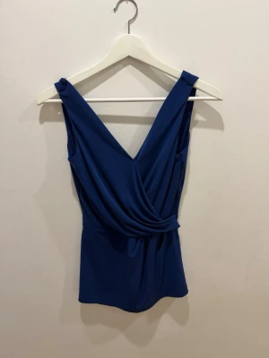 Mörkblå omlottopp med v-ringning - Snygg mörkblå omlottopp med breda axelband och djup v-ringning både fram och bak. Toppen har en draperad omlottdetalj framtill som ger en cool och trendig look. Tillverkad i ett mjukt och stretchigt syntetmaterial.