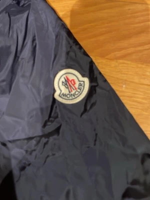 Mörkblå vindjacka från Moncler - Snygg mörkblå vindjacka från Moncler med huva och dragkedja framtill. Jackan har Moncler-logga på ärmen och praktiska fickor med knappar. Perfekt för blåsiga dagar och enkel att matcha med olika outfits.