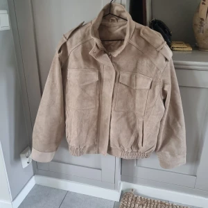 Zara Viral mockajacka helt ny - Snygg beige bomberjacka i mocka med två stora bröstfickor med lock, axeldetaljer och elastisk nederkant. Jackan har hög krage och stängs med dragkedja och tryckknappar. Perfekt för dig som gillar stilrena och coola jackor. Från samma koncern som Zara och Massimo Dutti som heter Grupo Inditex.