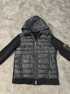 Moncler cardigan - Snygg svart dunjacka från Moncler med quiltad front och huva. Jackan har stickade ärmar och ribbade muddar, samt två fickor med tryckknappar och dragkedja framtill. Perfekt för kyliga dagar och riktigt stilren look.