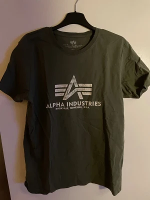 Mörkgrön Alpha Industries t-shirt - Snygg mörkgrön t-shirt från Alpha Industries med klassisk logga och text i vitt på bröstet. T-shirten har rund hals och är tillverkad i mjuk bomull som känns skön mot huden. Perfekt för en avslappnad och stilren look.