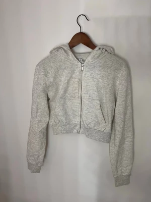 Ljusgrå croppad hoodie med dragkedja - Snygg ljusgrå croppad hoodie med dragkedja framtill och huva. Hoodien har långa ärmar, ribbade muddar och en stor ficka framtill. Perfekt för en chill och avslappnad stil.