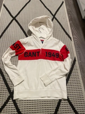 Vit hoodie från Gant med röd detalj - Snygg vit hoodie från Gant med bred röd rand över bröstet och armarna där det står 'GANT 1949' och 'CONNECTICUT'. Hoodien har huva, långa ärmar och ribbade muddar. Perfekt för en sportig och avslappnad stil.