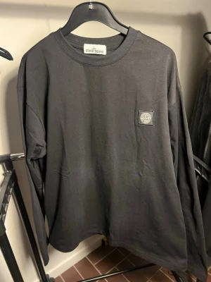  Stone Island  - Säljer en svart långärmad t-shirt från Stone Island med klassisk patch på bröstet. Tröjan har rund halsringning och är gjord i mjuk bomull för en skön och avslappnad stil. Perfekt för dig som gillar streetwear och vill ha något stilrent.
