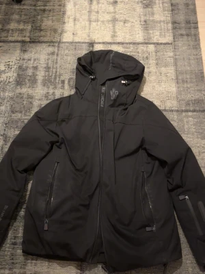 Svart dunjacka från Moncler - Snygg svart dunjacka från Moncler med huva och dragkedja framtill. Insidan har ett glansigt foder med Monclers ikoniska röd, vit och blå rand. Två fickor med dragkedja på utsidan och justerbar nederkant. Perfekt för kalla dagar.