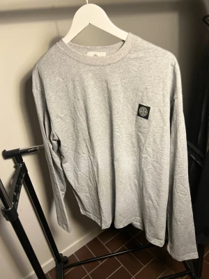  Stone Island - Snygg grå långärmad tröja från Stone Island med klassisk rund hals och ikonisk patch på bröstet. Tillverkad i mjuk bomull för skön känsla och enkel stil. Perfekt för dig som gillar streetwear och enkel design.